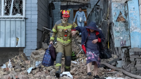 A firefighter helps an old lady out a building holding her arm as they step over piles of bricks and rubble