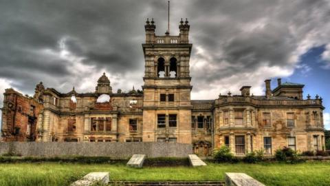 The main facade of Overstone Hall, a stone-built Victorian mansion with an Italianate tower in its middle. Its roof has largely gone, many of its windows lack glass or are boarded up. It has grass in front of it and there are stormy clouds above. 