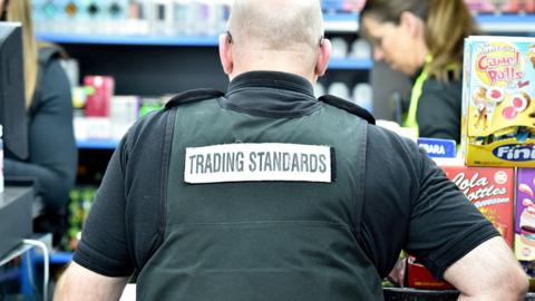 A trading standards officer in a shop with his back to the camera, wearing a black shirt and a stab vest with "trading standards" written across the back. Other officers are in the background.