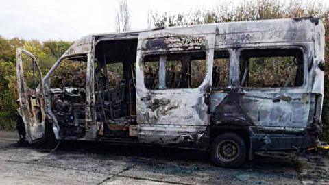 A burned out white minibus. It is parked on concrete with foliage in the background.