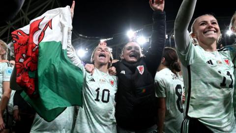 Jess Fishlock holds a Wales flag as she celebrates with Lily Woodham, Hannah Cain and other team-mates after their play-off win in Dublin