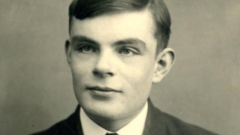 Alan Turing is seen in a black and white photo. He has sort hair is wearing a dark jacket, a white shirt and a tie