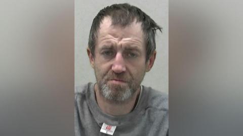 Mugshot of Christopher Scott. He has short scruffy dark hair and a beard and looks generally dishevelled. He has a morose expression on his face and is wearing a grey sweatshirt which is clearly inside out and back to front as the inner label is visible beneath his chin.