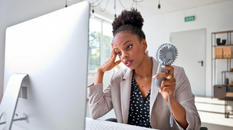 A woman sat in front of a computer screen, holding a mini fan, looking tired