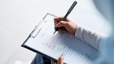 Two hands are shown holding a clipboard in the left hand and a pen in the right hand, about to write on the sheet. Some lines and boxes can be seen on the piece of paper.