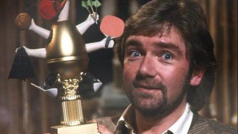 Noel Edmonds holding a Golden Egg award. An egg with multiple arms holding a pin pong bat, skies and other items and wearing skies.  Noel has a slightly surprised smile on his face.