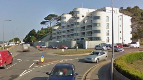 The roundabout on Inner Road has several cars on it and approaching it on a bright blue sky day. There is a large white building opposite it which has balconies and a rounded turret-style design.