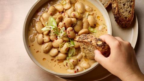 Creamy, saucy butter beans in a beige bowl with a hand dunking a chunk of bread