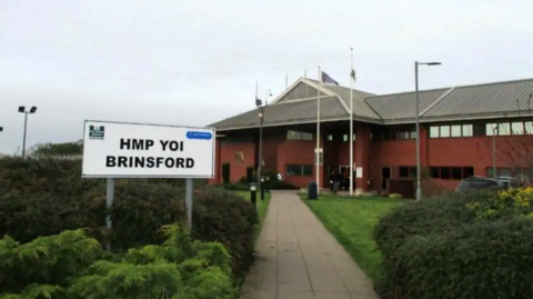 A sign including HMP YOI Brinsford in black letters in the foreground is near bushes. A building, including a grey roof at an angle to the wall for the first and ground floor, is in the background. 