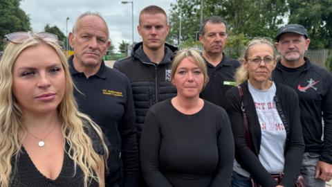 Seven business owners in a huddle, all wearing black standing in front of a tip with angry faces.