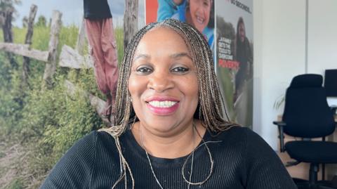 Fola Komolafe is seated indoors in an office setting. She has long braided hair and is wearing a long‑sleeved, ribbed black top. Behind her is a large printed backdrop featuring outdoor scenes with wooden fencing, greenery, and people standing in nature. To one side of the image, there is a standard office chair and a desk with a small plant on it. 