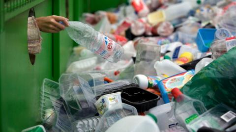 A hand is depositing a plastic Evian water bottle into a large green recycling bin. There are hundreds of plastic bottles and tubs in it