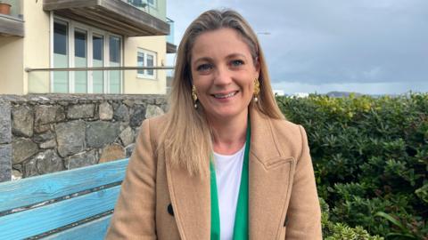 Ellie Lawrence is sitting outside on a blue wooden garden bench and she is smiling at the camera. She has long blonde hair and is wearing a white top, a bright green jacket, a pale brown coat and large gold and pearl earrings. Behind her is a building with French windows and a balcony and to her left are tall shrubs. In the far distance a part of a headland can be seen.