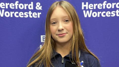 A young girl wearing a dark blue top stands in front of a purple banner decorated with the BBC Radio Hereford & Worcester logo. She is holding a black golf trophy which reads "Daily Mail World Golf Championship - Winner 2025."