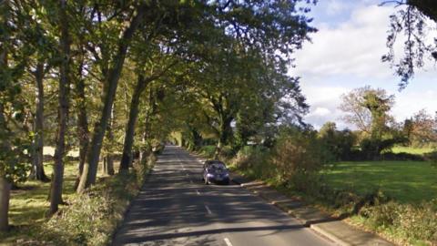 A google maps picture of the road in question which is to close. Trees line the side of the road whilst a car drives towards the camera.