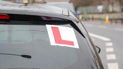 The back of a car on a road. It has a red L stuck to the window.