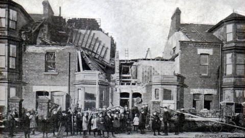 A black and white photograph of people trying to clear up rubble on Cleveland Road in Hartlepool after the bombardment. There is a house in the middle with its roof torn off and another one on the left with a collapsed  roof.