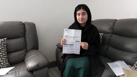 Arooj Hussain holds up a letter to the camera. She is sat on a grey leather sofa and is wearing a black headscarf and a green dress.