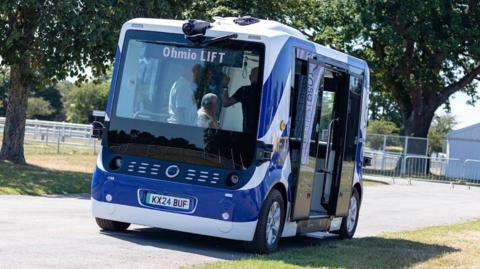A stationary blue and white self-driving bus. Its side doors are open and people are visible through its windscreen aboard the vehicle on a bright and sunny day.