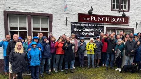 A mixed gender group of people in raincoats stand outside the Punch Bowl inn, raising glasses.