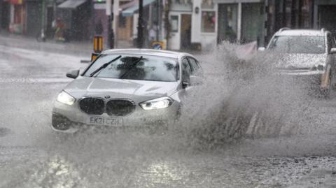 A silver car drives through water on the road in suburban London, creating a large splash. 