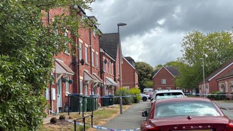 A residential road with cars parked on it. There is a police cordon on the street