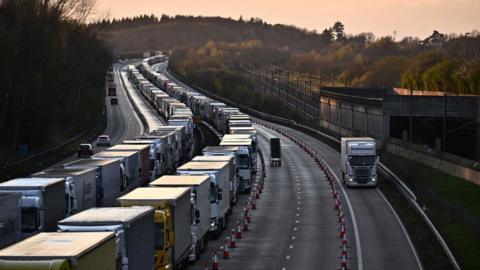 Lorries queue towards the camera in two lanes and in a queue that stretches around the corner and out of shot. On the far left of the image, traffic can be seen on the other carriageway travelling in the other direction. In the distance there are trees and an orange sunset.