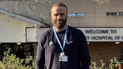 Jamie Roberts stood out the UHW Hospital in Cardiff. He is smiling wearing a navy blue jumper with his lanyard and stethoscope around his neck.