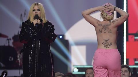 Avril Lavigne onstage at the Juno Awards, next to a topless protestor