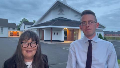 Martina Burke and Josiah Burke standing outside the Hebron Free Presbyterian Church in Ballymoney. Martina has long, dark, greying hair with a fringe, wearing glasses, a black blazer and white top. Josiah has short dark hair, wearing glasses, a white shirt and dark tie. The church is in the background and has a triangular roof.