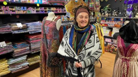Kim Roberts, who has brown hair tied into dreadlocks and wears a black and white shawl and a colourful scard, smiles at the camera in a clothes shop.