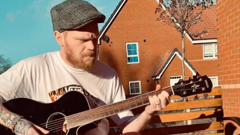 Craig Brown, wearing a flat cap and t-shirt, sits on a bench outside while playing guitar.