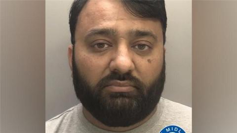 A man with short black hair and a black beard looks at the camera in a police custody photograph.