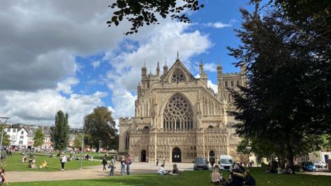 A view of Exeter Cathedral from Cathedral Green with a tree in the foreground on the left and people sitting out on the green