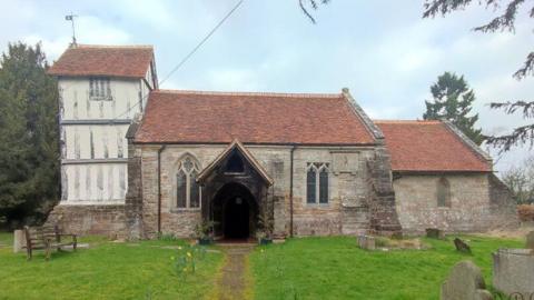A church in a church yard with green grass in front of it. The doorway of the church is darkened by damage. 