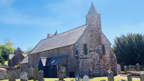 Church building - Arbuthnott Church - with graves in the foreground, under a blue sky.