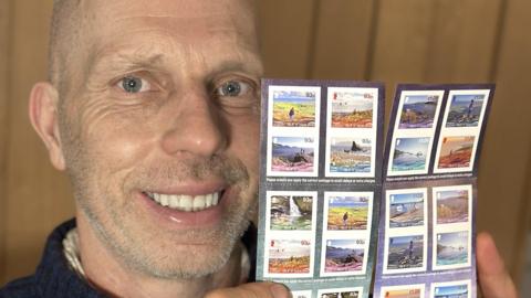 Andy North holding the stamp collection booklets. A bald man, he os wearing a navy blue jumper.