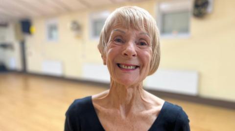 Barbara Wraith is standing in a hall. She has short blonde hair and is wearing pink lipstick and a black V-neck T-shirt.