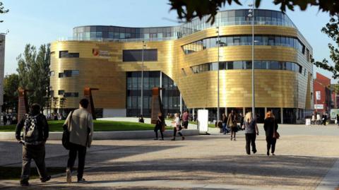 Teesside University. It is a modern, curved building, made of gold-coloured panels and glass. There are several people walking around. 