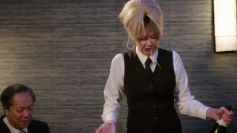 Mandy, in a waitress outfit, holds an open bottle of champagne while looking stressed