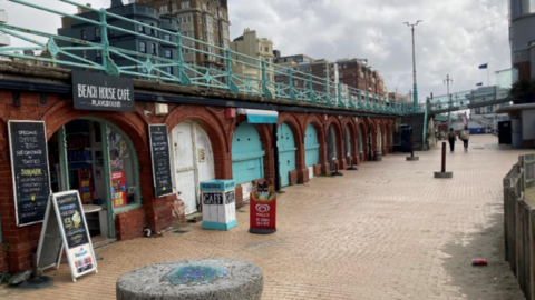 Red brick arches under Brighton promenade. There are shops in some of the arches and light blue handrails on the promenade.