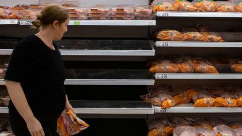 A woman dressed all in black looks at chicken on display in a supermarket fridge. Some of the shelves are empty.
