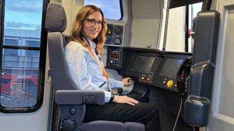 A woman sat at the helm of a train. She has hair just below her shoulders and is wearing glasses, a blue shirt and black trousers. In the train's cab are various screens, levers and and dials.