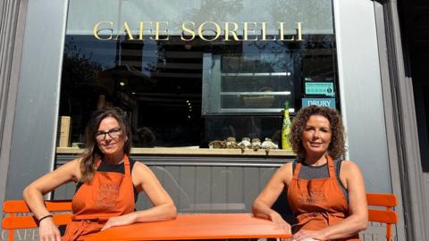 Serafina Zambito with long straight brown hair and glasses, and Jo Garvin with curly brown hair, both wearing orange aprons and sitting in orange chairs either side of an orange table. There is a silver-framed window behind with CAFE SORELLI in yellow letters towards the top of the window.