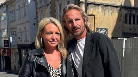 Nigel Adams wearing a white T-shirt and black blazer. He has long blonde hair and a dark beard. He is standing beside his fiancee Louise Payne, who has blonde hair and is wearing large hoop earrings, a cheetah print blouse and a black leather jacket.