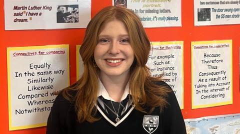 Imogen is looking at the camera and wearing a dark blue school uniform with a white shirt ands silver crest. she has chest lenght wavy red hair and has a broad smile showing her teeth