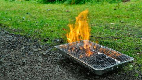 A disposable barbecue straddles a soily patch and grass as it burns.