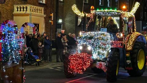 A tractor is brightly-lit with reindeer antlers and a red nose and driving through Oswestry. People are standing on the pavements watching and the tail end of another vehicle with fairy lights can be seen. The tractors are passing a building with a decorative entrance.