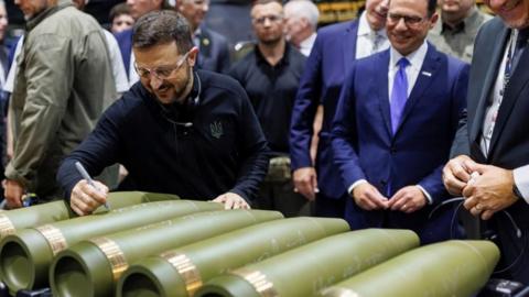 Ukrainian President Volodymyr Zelensky smiles as he writes on a shell at an American munitions factory 