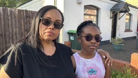 Angela with her 11-year-old daughter Ava. They are both wearing sunglasses on a pleasant day. Angela has long black hair, black sunglasses and a black top on. Ava has black hair tied up, black sunglasses and is wearing a pink vest top with a Disney character on.